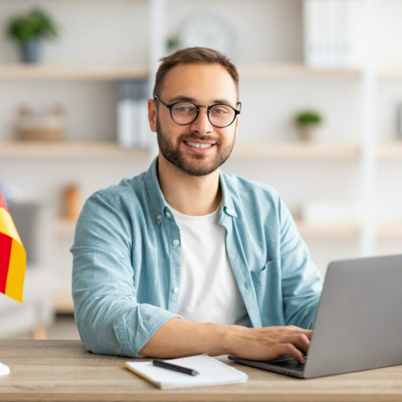 Happy young guy sitting at table with flag of Germany, using laptop pc, studying foreign language