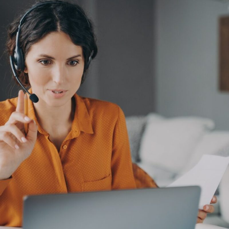 Young Spanish woman tutor wearing headset looking at laptop screen during online class